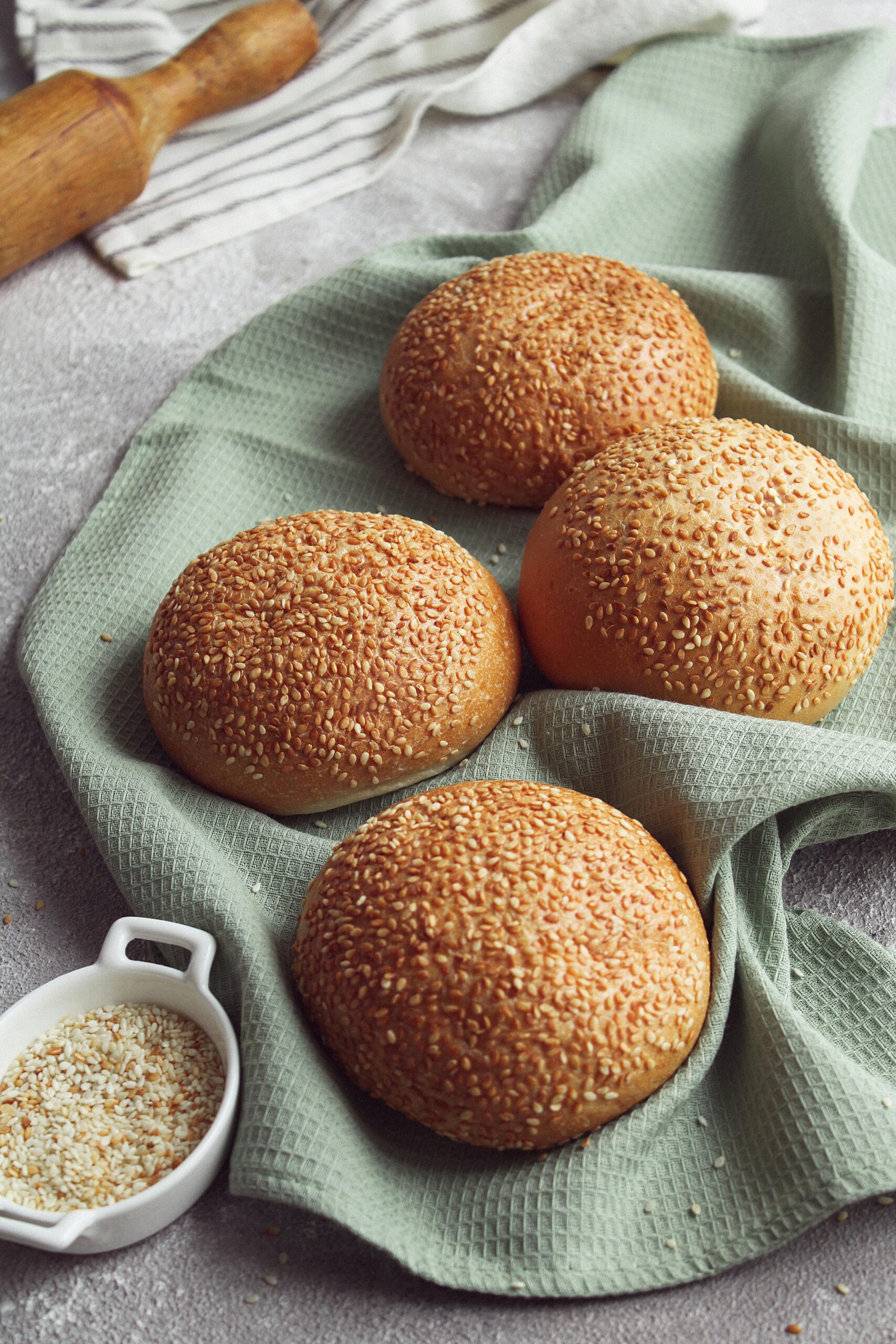 Close-up of four sesame seed buns displayed on a textured green cloth with a sprinkle of seeds nearby.